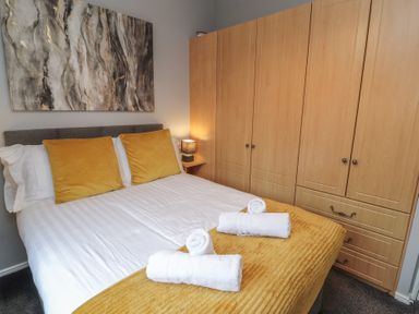 A bedroom with a double bed with yellow cushions and blanket wooden wardrobes and a bedside table with a lamp at Lakeside 18 in Carnforth
