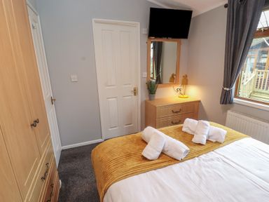 A bedroom with a bed with rolled towels on a yellow blanket a wooden dresser with a lamp and plant a mirror and a TV on the wall at Lakeside 18 in Carnforth