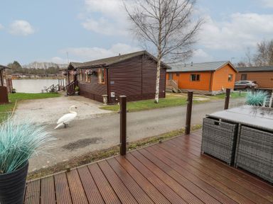 A wooden deck with outdoor furniture overlooking a road with cottages and a swan near a lake at Lakeside 18 in Carnforth