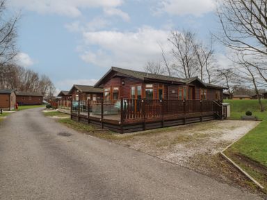 A wooden cabin with a fenced deck next to a paved road and trees at Lakeside 18 in Carnforth