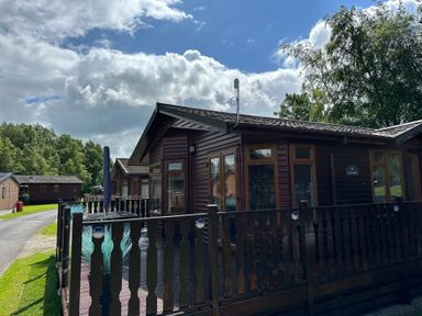 A wooden cabin with a fenced deck and outdoor furniture under a partly cloudy sky at Lakeside 18 in Carnforth
