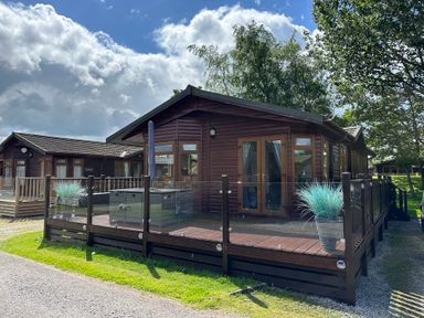 A wooden cabin with a deck and glass railing surrounded by grass and trees at Lakeside 18 in Carnforth