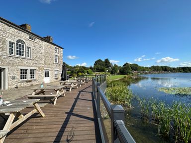 A stone building with wooden picnic tables on a deck by a lake with grass and trees at Lakeside 18 in Carnforth