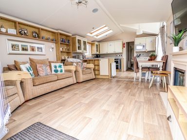 An open plan living area with a beige sofa and armchair a dining table with chairs a kitchen with cabinets and appliances and wooden flooring at Hazel Park Lodge HP4 Rhyl