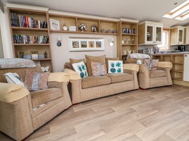 A living room with three beige armchairs, cushions with floral patterns, built-in bookshelves with books and decorative items, and a kitchen area in the background at Hazel Park Lodge HP4 in Rhyl