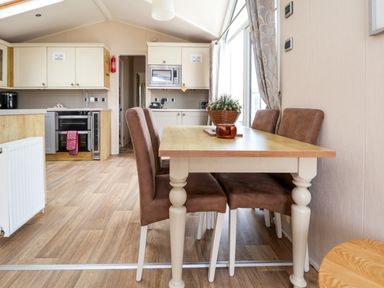 A dining area with a wooden table and four brown chairs next to a kitchen with white cabinets at Hazel Park Lodge HP4 in Rhyl