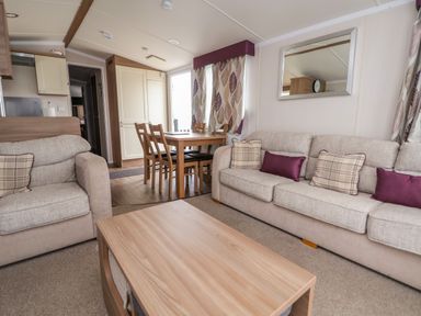 A living room with beige sofas, a wooden coffee table, dining table with chairs near a window and patterned curtains at Hazel Park 11 HP11 in Rhyl