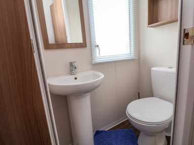 A small bathroom with a white pedestal sink mirror window with blinds white toilet and blue bath mat at Hazel Park 11 HP11 in Rhyl