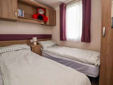 A bedroom with two single beds a bedside table and lamp a window with curtains and overhead storage cabinets at Hazel Park 11 HP11 in Rhyl