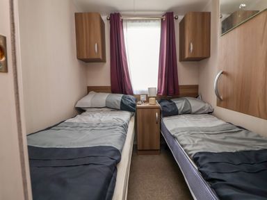 A bedroom with two single beds separated by a wooden nightstand with a lamp and remote control and purple curtains on the window at Hazel Park 11 HP11 in Rhyl