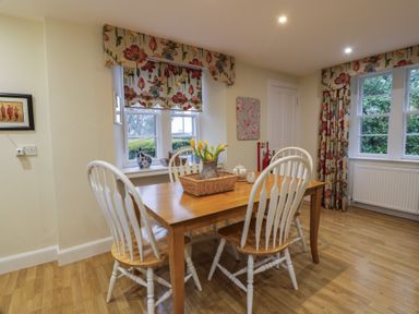 A dining area with a wooden table and white chairs with floral curtains and windows at Eagle Glen Cottage in Kettins near Coupar Angus