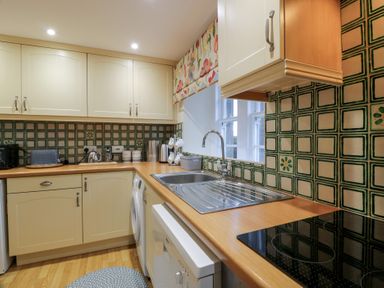 A kitchen with cream cabinets a stainless steel sink a washing machine a ceramic cooktop and patterned green tiles on walls at Eagle Glen Cottage in Kettins near Coupar Angus