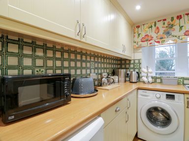 A kitchen with microwave toaster kettle washing machine and floral curtain over window at Eagle Glen Cottage in Kettins near Coupar Angus