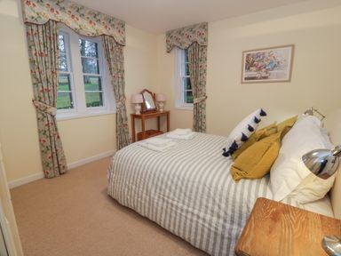 A bedroom with a bed with striped bedding cushions and lamps near windows with floral curtains at Eagle Glen Cottage in Kettins near Coupar Angus