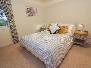 A bedroom with a double bed striped bedding and towels on top two bedside tables with lamps a window with floral curtains and a framed picture on the wall at Eagle Glen Cottage in Kettins near Coupar Angus