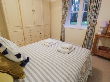 A bedroom with a bed striped bedding and folded towels a wardrobe window with floral curtains and a wooden side table with a lamp at Eagle Glen Cottage in Kettins near Coupar Angus