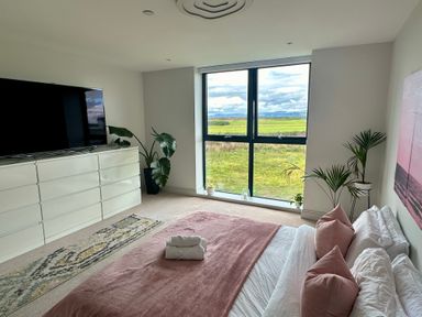 A bedroom with a large window showing grass and hills a bed with pink pillows and blanket a white dresser with a television and plants at The Links 40 An apartment with spectacular sea views Porthcawl