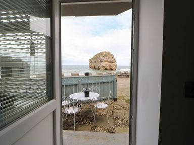 View through an open door showing a small outdoor area with a white metal table and three chairs overlooking the sea and a rock formation at The Beach Apartment in South Shields
