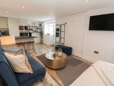A living area with a blue sofa, round coffee table, bar stools, kitchen, full-length mirror, and wall-mounted TV at The Beach Apartment in South Shields