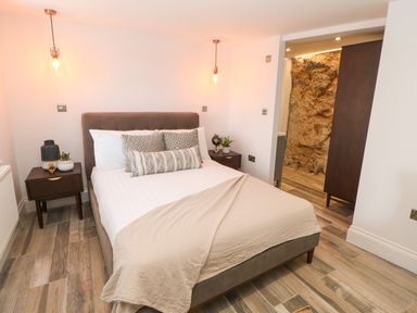 A bedroom with a bed and patterned pillows two bedside tables with lamps and plants wooden flooring and an exposed rock wall at The Beach Apartment in South Shields