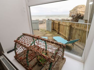 A window view showing a fenced patio with two blue lounge chairs and a picnic table near the sea with a rock formation at The Beach Apartment in South Shields