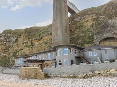 A building with a tall chimney-like structure set against rocky cliffs by a pebble beach at The Beach Apartment in South Shields