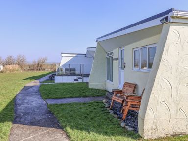 Outdoor area with a concrete walkway grass patches and a small patio with two wooden chairs at Atlantic Escape in Widemouth Bay