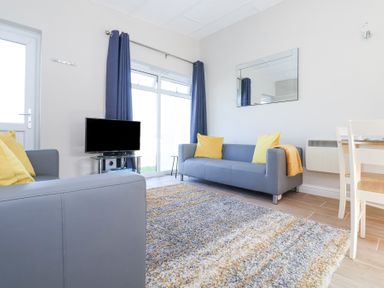 A living room with gray sofas yellow cushions a television on a stand a rug and a dining table at Atlantic Escape in Widemouth Bay