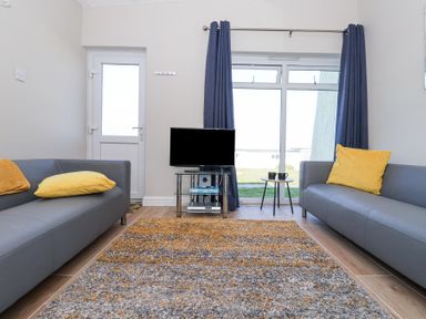 A living room with two gray sofas with yellow cushions a television on a glass stand a small round table with two mugs and blue curtains at Atlantic Escape in Widemouth Bay