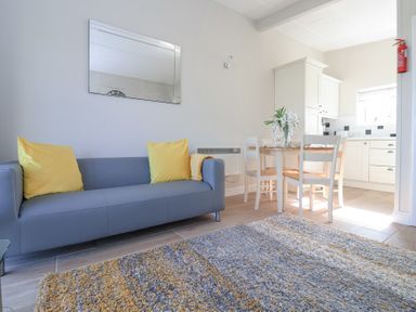 A living area with a grey sofa with yellow cushions a dining table with white chairs and a kitchen in the background at Atlantic Escape in Widemouth Bay