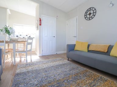 A living room with a gray sofa with yellow cushions a patterned rug a dining table with chairs and a kitchen area at Atlantic Escape in Widemouth Bay