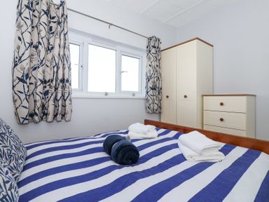 A bedroom with a striped bedspread a pillow folded towels floral curtains and cream wardrobe and chest of drawers at Atlantic Escape in Widemouth Bay