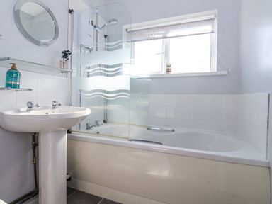 A bathroom with a sink a bathtub with shower and frosted glass screen a window and a round mirror at Atlantic Escape in Widemouth Bay