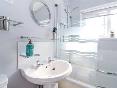 A bathroom with a white sink a round mirror a bottle of soap and a bathtub with a glass shower screen at Atlantic Escape in Widemouth Bay