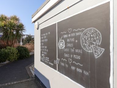 A chalkboard menu on an exterior wall with drawings of food and opening times beside a path with plants at Atlantic Escape in Widemouth Bay