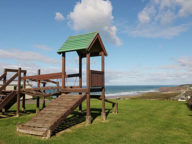 A wooden playground structure on grass with a view of the ocean and cliffs at Atlantic Escape in Widemouth Bay
