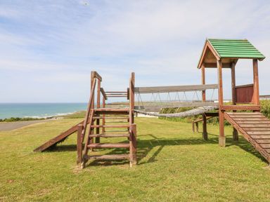 A wooden playground structure on grass with the ocean in the background at Atlantic Escape in Widemouth Bay