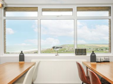A dining area with wooden tables and chairs by a large window overlooking a coastal landscape at Atlantic Escape in Widemouth Bay
