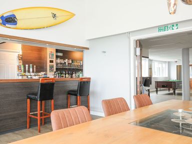 A bar area with two black and wood bar stools a yellow surfboard mounted on the wall and a wooden table with brown chairs in a room with a pool table beyond the doorway at Atlantic Escape in Widemouth Bay