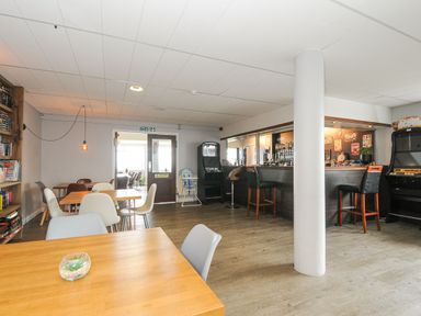 A bar area with tables, chairs, a bookshelf, arcade machines, and a counter with bar stools at Atlantic Escape in Widemouth Bay