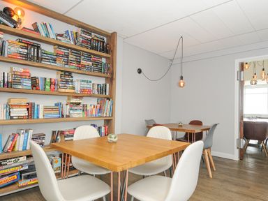 A room with wooden tables white and gray chairs a wooden bookshelf filled with books and board games and hanging light bulbs at Atlantic Escape in Widemouth Bay