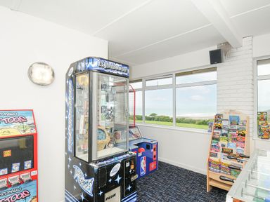 A game room with arcade machines a brochure stand and large windows showing a grassy area and ocean at Atlantic Escape in Widemouth Bay