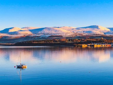 A lake with a sailboat and distant snow-covered hills under a clear sky at Cwt Mochyn in Llanberis