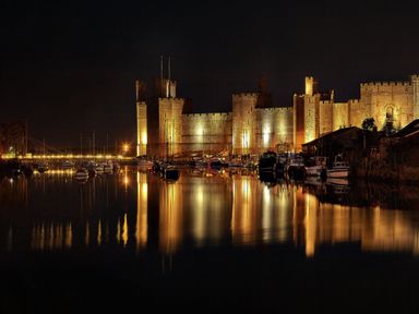 A castle illuminated at night with boats docked in calm water reflecting the lights