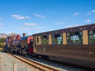A steam locomotive pulling a passenger carriage on railway tracks with mountains in the background at Cwt Mochyn in Llanberis