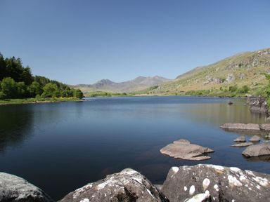 A lake surrounded by rocks trees and hills with mountains in the background at Cwt Mochyn in Llanberis