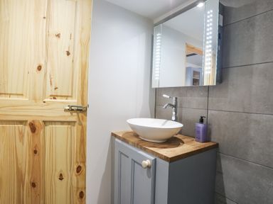 A bathroom sink with a round white basin on a wooden countertop and a mirror with lights above at Cwt Mochyn in Llanberis