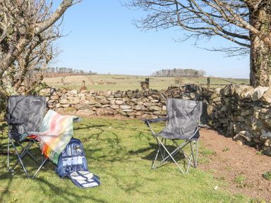 Two folding camping chairs on grass near stone wall and trees with a picnic set and blanket at Cwt Mochyn in Llanberis
