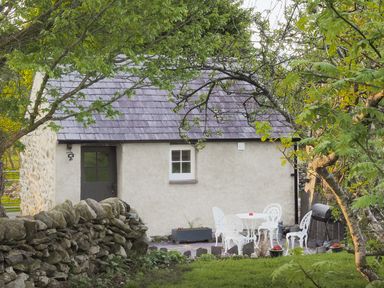 An outdoor area with white metal chairs and table near a stone wall and cottage at Cwt Mochyn in Llanberis