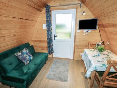 A small wooden room with a green sofa a floral pillow a door a wall-mounted tv a table with a floral tablecloth and chairs with a plant and a wine bucket Chantelles Holbeach